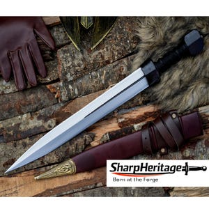 Könnte beinhalten: Ein silbernes und schwarzes Schwert mit lederummanteltem Griff und einer braunen Lederscheide. Das Schwert liegt auf einer Holzoberfläche mit einem braunen Lederhandschuh und einem gold-schwarzen Helm im Hintergrund. Der Text "SharpHeritage Born at the Forge" ist sichtbar.