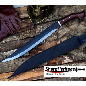 May include: A large, bladed knife with a dark wooden handle and a black sheath. The blade is silver and the handle is a dark brown with silver rivets. The knife is displayed on a wooden surface with a leather glove and a gold helmet in the background. The text "SharpHeritage" is visible.
