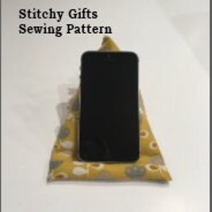 May include: A yellow and white patterned fabric phone stand. The stand is shaped like a triangle and is filled with stuffing. A black smartphone is resting on the stand.