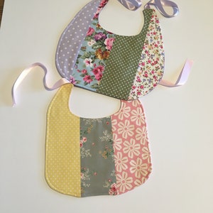 Peut inclure: Deux bavoirs pour bébé faits à la main avec différents motifs floraux. Les bavoirs sont en tissu de coton et ont un motif de pois rose clair et blanc sur le dessus. Les bavoirs ont un ruban rose clair attaché à eux.