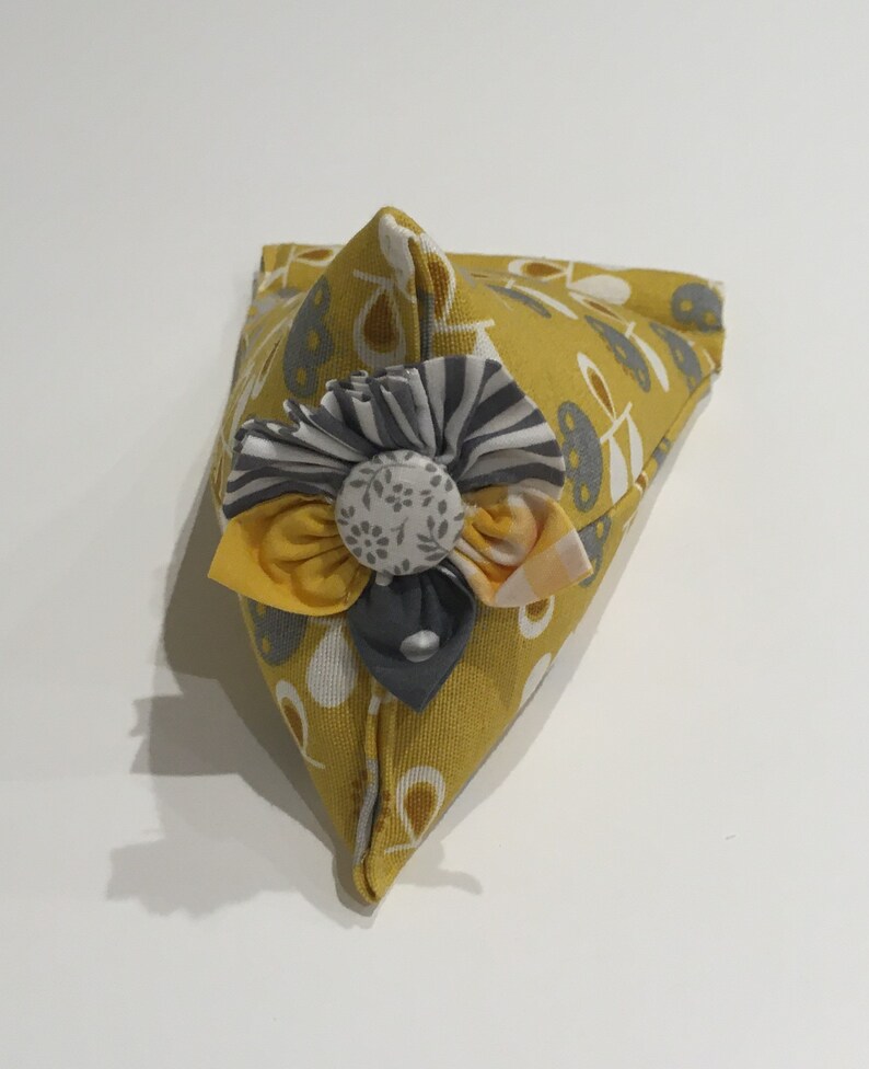 May include: A yellow triangular pincushion with a white and gray floral pattern. The pincushion has a gray and white fabric flower with a white button in the center.