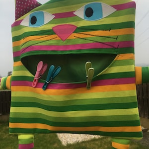 May include: A whimsical, striped cat-shaped laundry bag in shades of green, pink, and orange. The bag features large blue eyes, a pink nose, and a wide mouth. Three colorful clothespins are visible inside the open mouth. The cat has arms, legs, and ears with pink polka dots.