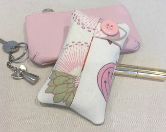 Pode incluir: Um pequeno porta-lenços de tecido com padrão floral rosa e verde com fecho de botão rosa. O porta-lenços está sobre uma superfície bege claro.