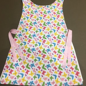 May include: A white apron with a butterfly print in shades of pink, yellow, blue, green, and purple. The apron has two pink ties.