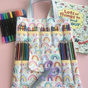 Könnte beinhalten: Eine weiße Stofftasche mit einem Regenbogenmuster und einer Tasche auf der Vorderseite. Die Tasche hat einen Reißverschluss und einen Griff. Die Tasche ist gefüllt mit Buntstiften, Farbstiften und einer Schere. Die Tasche liegt auf einer rosa Oberfläche. Der Text auf dem Buch lautet "Viele Dinge zum Ausmalen".