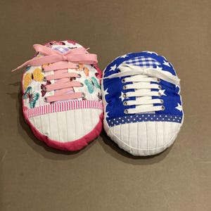 May include: Two fabric shoe-shaped pillows. One is pink with a butterfly pattern and the other is blue with white stars. Both have white laces and a white toe.
