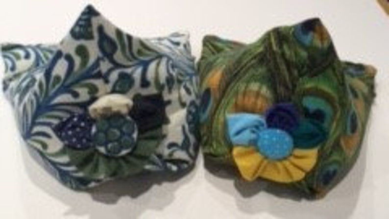 May include: Two fabric face masks with floral patterns. One mask is green and white with a blue button, the other is green with blue, yellow, and white fabric accents.