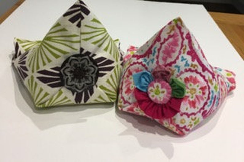 May include: Two fabric bookends, one with a green and brown floral pattern and the other with a pink and white floral pattern. Both bookends have a triangular shape and are decorated with a flower.