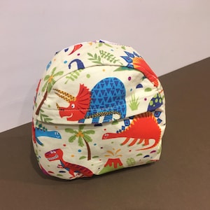 May include: A small, cream-colored backpack with a dinosaur print. The backpack features a variety of dinosaurs, including a triceratops, a stegosaurus, and a tyrannosaurus rex. The dinosaurs are depicted in bright colors, including red, blue, green, and yellow.