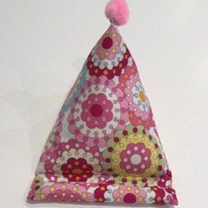 May include: A pink and white floral patterned pillow with a pink pom-pom on top. The pillow is shaped like a triangle and is designed to prop up a book or tablet.