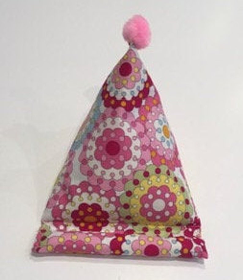 May include: A pink and white floral patterned pillow with a pink pom-pom on top. The pillow is shaped like a triangle and is designed to prop up a book or tablet.