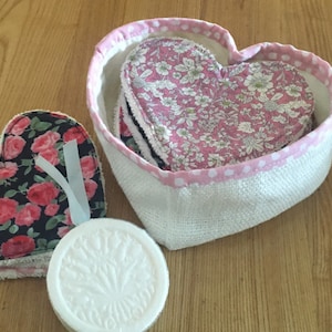 May include: A set of heart-shaped reusable makeup remover pads in a pink and white heart-shaped container. The pads are made of fabric with floral patterns and are stacked on top of each other. There is a white round soap bar with a floral design on the surface.