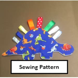 May include: Blue fabric dinosaur-shaped baby toy with a variety of colorful ribbons attached. The fabric features a repeating pattern of dinosaurs in various colors.