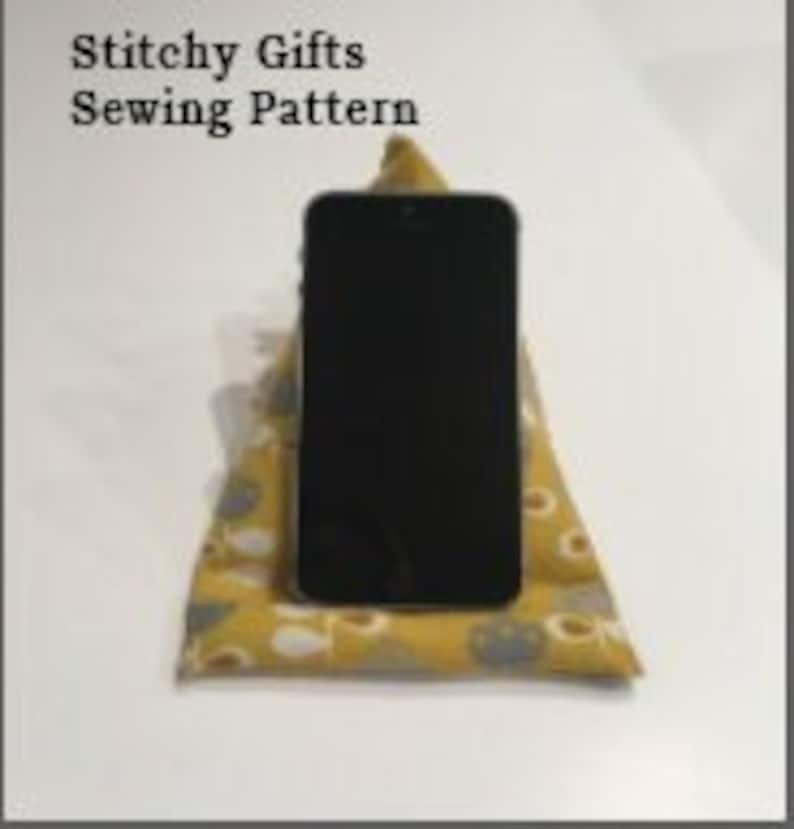 May include: A yellow and white patterned fabric phone stand. The stand is shaped like a triangle and is filled with stuffing. A black smartphone is resting on the stand.