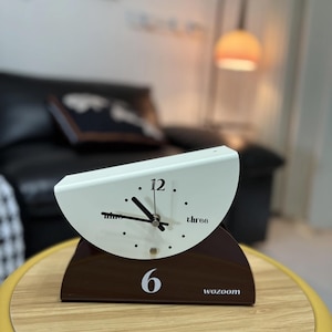 May include: A modern, minimalist clock with a white face and a brown base. The clock face displays the numbers 12, 3, 6, and 9, with the word "wozoom" printed on the base. The clock is on a wooden table.