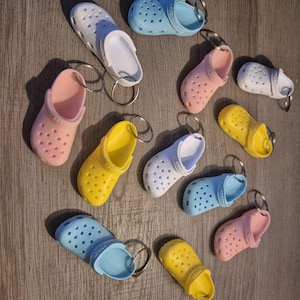 Può includere: Una collezione di portachiavi Crocs in miniatura in vari colori, tra cui bianco, rosa, giallo e blu. Ogni portachiavi presenta una piccola e dettagliata replica del popolare design della scarpa, completa di anello in metallo.