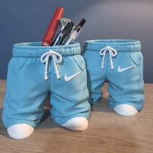 Può includere: Due contenitori decorativi azzurri a forma di pantaloni della tuta, con coulisse bianche e un logo Nike bianco. Un contenitore contiene pennelli per il trucco e un rossetto rosso. I contenitori hanno basi bianche a forma di scarpa.