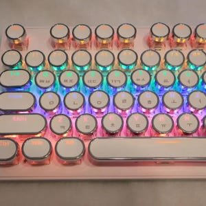 Puede incluir: Primer plano de un teclado mecánico con teclas redondas blancas y una base rosa. Las teclas están retroiluminadas con varios colores, incluyendo azul, verde y rojo. El teclado tiene una estética retro.