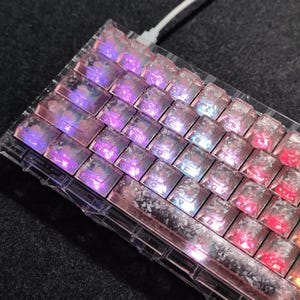May include: A close-up of a clear, custom mechanical keyboard with iridescent keycaps. The keycaps have a pink and purple color scheme, with a clear, textured appearance. A white cable is plugged into the keyboard.