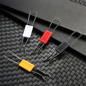 Könnte beinhalten: Vier Tastenkappen-Abzieher in Weiß, Rot, Schwarz und Gelb. Jedes Werkzeug hat einen rechteckigen Griff und Drahtzinken zum Entfernen von Tastenkappen von mechanischen Tastaturen. Die Werkzeuge sind auf einer strukturierten schwarzen Oberfläche angeordnet.