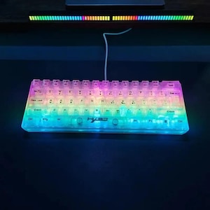 Könnte beinhalten: Eine mechanische Tastatur mit Regenbogen-Hintergrundbeleuchtung und transparenten Tasten. Die Tastatur ist über ein weißes Kabel verbunden. Oberhalb der Tastatur befindet sich eine passende LED-Lichtleiste. Standard-QWERTZ-Layout.