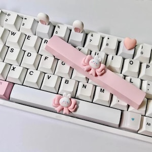 Può includere: Una tastiera meccanica bianca con dettagli rosa. La tastiera presenta una barra spaziatrice rosa con un dettaglio a forma di coniglietto e fiocco, tasti a forma di orecchie di coniglio e un tasto a forma di cuore. Un oggetto decorativo.