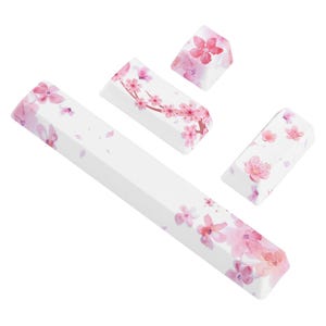 May include: White keyboard keycaps with a pink cherry blossom design. The set includes a long space bar keycap, and three other keycaps with varying floral patterns. The blossoms are in shades of pink and red.