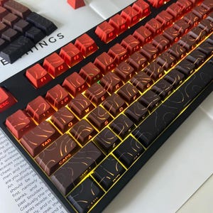 May include: A mechanical keyboard with a black frame and keycaps in shades of red, brown, and black. The keycaps have a unique swirl design and are backlit with a warm glow. Additional keycaps are visible in the background.