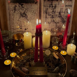 May include: A dimly lit scene featuring candles, a brass tray with herbs, and two framed illustrations. Tall red candles are lit, along with smaller tea lights. The brass tray has a sign that reads "Sacred Lineage Rituals".