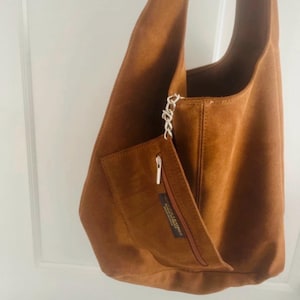 May include: A brown suede shoulder bag with a large main compartment and a smaller front pocket. The bag has a long strap and a silver chain detail. The front pocket has a zipper closure. The bag is made of a soft, suede-like material.