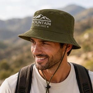 以下が含まれることがあります： 白の刺繍による山のグラフィックと「MOUNTAIN YOUR TEXT」の文字が施されたオリーブグリーンのバケットハット。同色のあご紐と調節可能なトグル、下向きのつば、側面の通気用アイレットが特徴です。