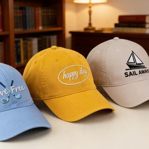 Puede incluir: Tres gorras de béisbol. Una gorra azul claro con "Live Free" y un gráfico de palo de golf, una gorra amarilla con el texto "happy day" y una gorra beige con un velero y el texto "SAIL AWAY".