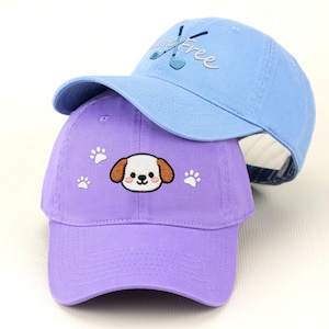 May include: Two baseball caps, one light blue and one purple. The light blue cap has the word "Free" embroidered on it, along with golf clubs. The purple cap features a puppy face and paw prints.