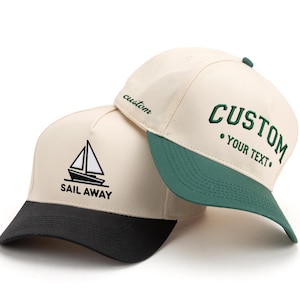 Puede incluir: Dos gorras de béisbol con coronas color crema. Una gorra tiene una visera negra y un gráfico de velero negro con el texto SAIL AWAY. La otra gorra tiene una visera verde y texto bordado en verde CUSTOM YOUR TEXT con custom en el lateral.