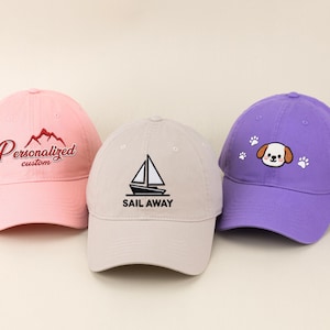 May include: Three baseball caps in pink, tan, and purple. The pink cap has "Personalized custom" embroidered in red. The tan cap has a sailboat graphic and "SAIL AWAY" in black. The purple cap features a dog face and paw prints.