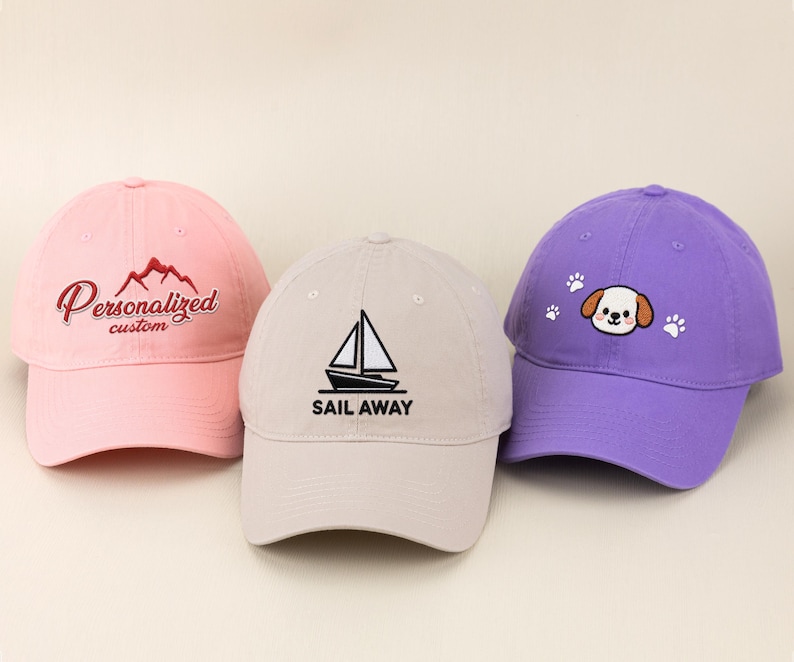 May include: Three baseball caps in pink, tan, and purple. The pink cap has "Personalized custom" embroidered in red. The tan cap has a sailboat graphic and "SAIL AWAY" in black. The purple cap features a dog face and paw prints.