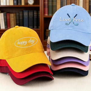 May include: A stack of baseball caps in various colors, including yellow, red, and blue. The yellow cap has the text "happy day" in white script. The blue cap features crossed golf clubs and the words "Live Free".