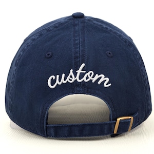 May include: Navy blue baseball cap with the word "custom" embroidered in white script across the back. The cap has a curved brim, a brass-colored adjustable buckle, and a button on top.