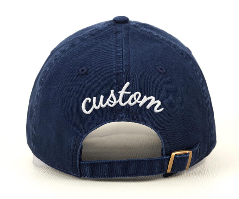 May include: Navy blue baseball cap with the word "custom" embroidered in white script across the back. The cap has a curved brim, a brass-colored adjustable buckle, and a button on top.