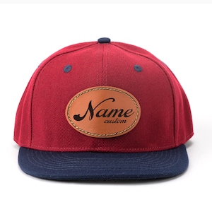 Op de afbeelding: Bordeauxkleurige baseballcap met een marineblauwe klep. De pet heeft een bruin leren label met het woord "Name" in een scriptlettertype en het woord "custom" eronder.