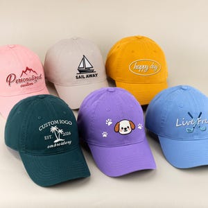 May include: A collection of six baseball caps in various colors, including pink, green, yellow, purple, and blue. Each cap features embroidered designs and text, such as a sailboat, a mountain, and the words "Happy Day" and "Live Free".