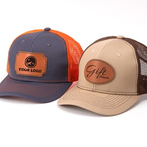 Pode incluir: Duas bonés de caminhoneiro com apliques de couro. Uma é azul marinho com costas de malha laranja e o texto "YOUR LOGO". A outra é bege com costas de malha marrom e o texto "Gift Keepsake".