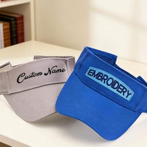 May include: Two visors are shown, one grey and one blue. The grey visor has "Custom Name" embroidered in black script. The blue visor features "EMBROIDERY" in a rectangular patch. Both visors are designed to shield the eyes from the sun.