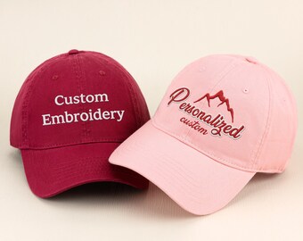 May include: Two baseball caps. One is burgundy with "Custom Embroidery" in white. The other is pink with "Personalized custom" and a mountain design in red. Both have curved visors.