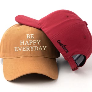 以下が含まれることがあります： 白の刺繍文字「BE HAPPY EVERYDAY」が入ったタンカラーの野球帽と、側面に黒の刺繍文字「custom」が入ったマルーンカラーの野球帽。どちらの帽子もカーブしたつば、構造化されたクラウン、調節可能なバックストラップを備えています。