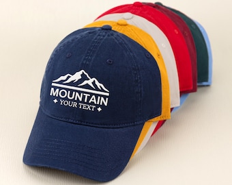 May include: A collection of baseball caps in various colors, including navy blue, yellow, red, and green. The navy blue cap in the front features the text "MOUNTAIN YOUR TEXT" in white, along with a mountain graphic. The caps are arranged in a row.