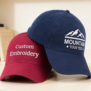 May include: Two baseball caps, one burgundy and one navy blue. The burgundy cap has "Custom Embroidery" in white script. The navy cap features a mountain graphic with the word "MOUNTAIN" and space for custom text in white.