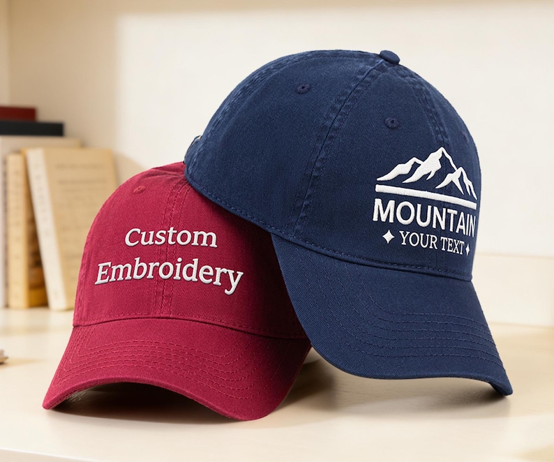 May include: Two baseball caps, one burgundy and one navy blue. The burgundy cap has "Custom Embroidery" in white script. The navy cap features a mountain graphic with the word "MOUNTAIN" and space for custom text in white.