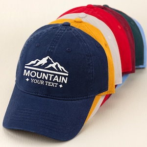 May include: A row of baseball caps in various colors, including navy blue, yellow, red, and green. The navy blue cap in the front features a white mountain graphic with the text "MOUNTAIN YOUR TEXT".