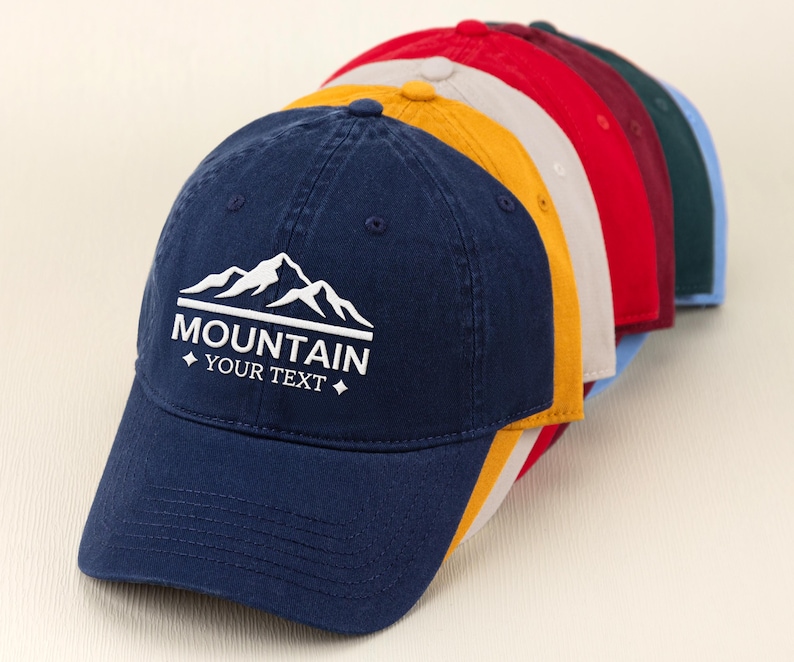 May include: A row of baseball caps in various colors, including navy blue, yellow, red, and green. The navy blue cap in the front features a white mountain graphic with the text "MOUNTAIN YOUR TEXT".